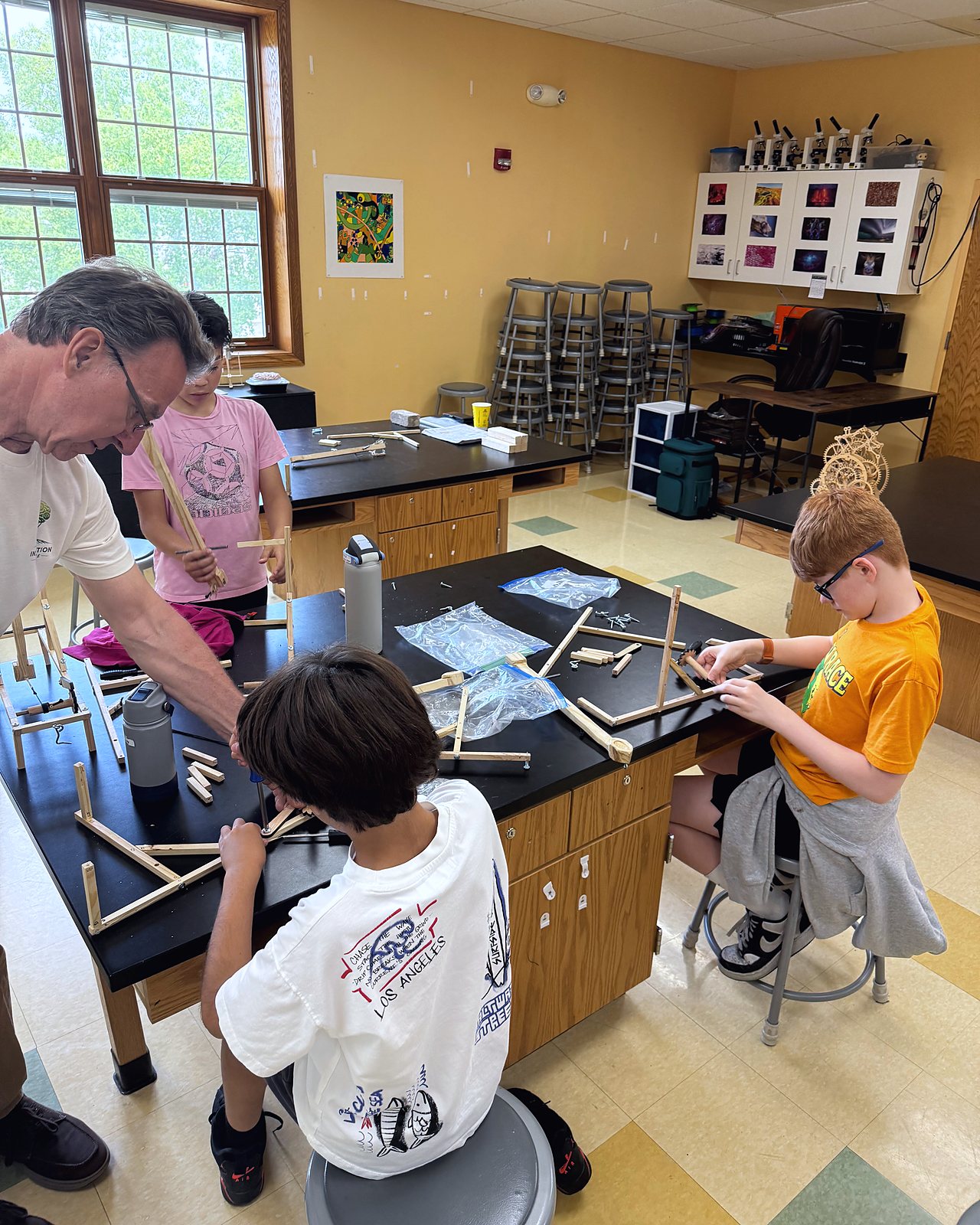 A guest instructor helps two students fit the arm of their catapult at the lab bench