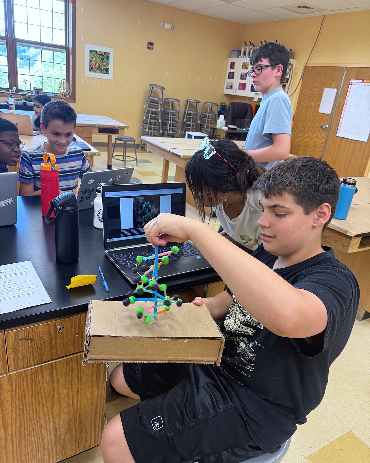 A student holds up the DNA helix he built over its cardboard base, his design open on the laptop behind him