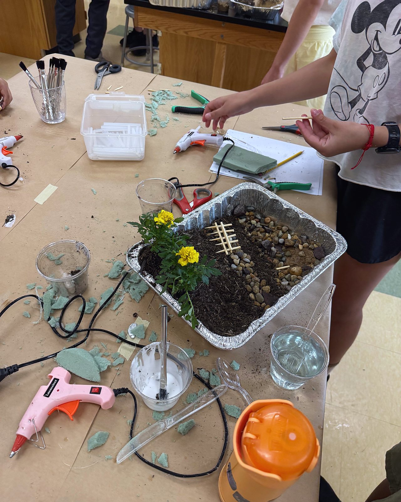 The hillside model taking shape in a foil tray: soil, stones, a fence of sticks and marigolds, with glue guns on the table