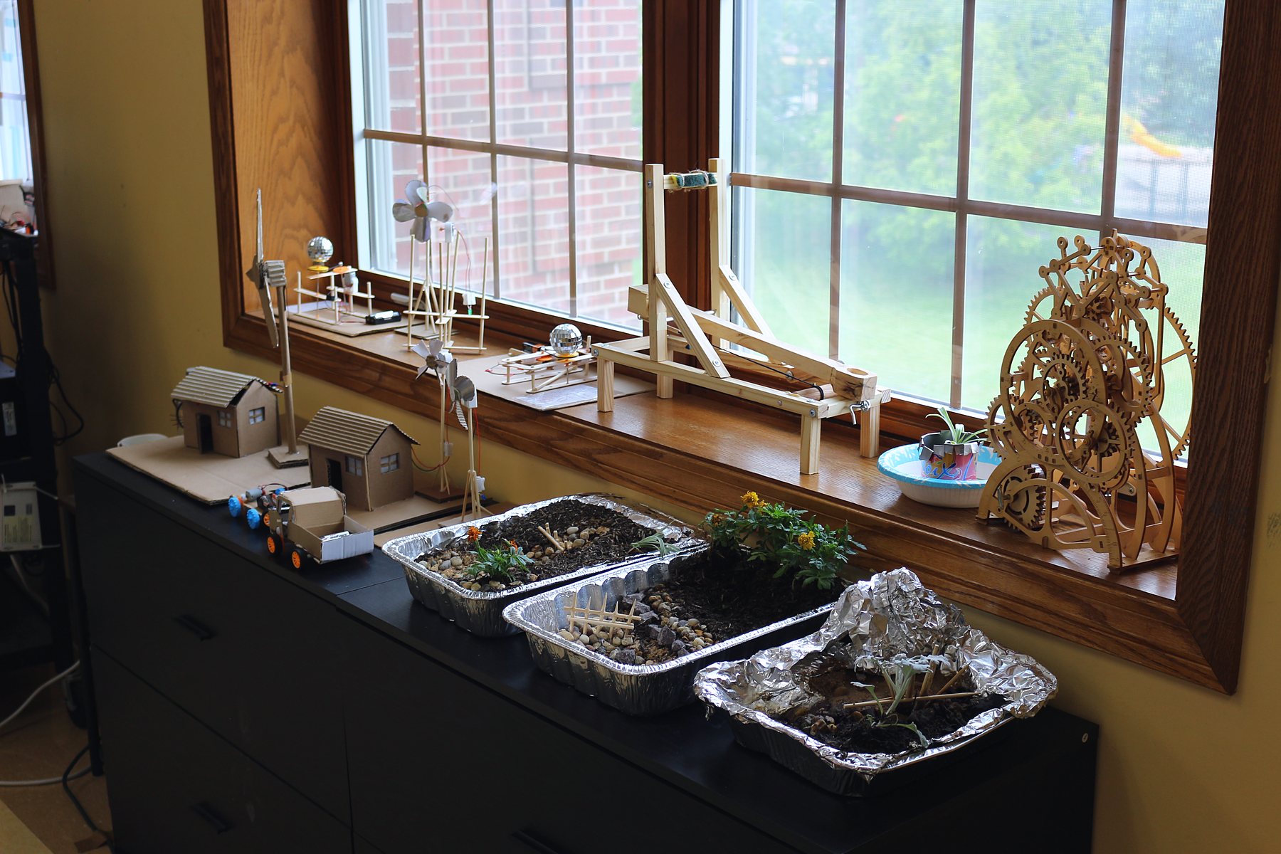 Student builds on the classroom windowsill: houses, turbines, a catapult, a wooden gear clock and habitat trays